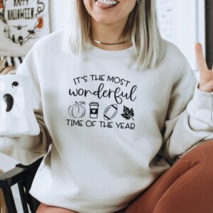 Puede incluir: Sudadera color crema con el texto "It's the most wonderful time of the year" y gr&aacute;ficos tem&aacute;ticos de oto&ntilde;o. El dise&ntilde;o incluye una calabaza, una taza de caf&eacute;, un bal&oacute;n de f&uacute;tbol americano y una hoja. Se sostiene una taza blanca con forma de fantasma.