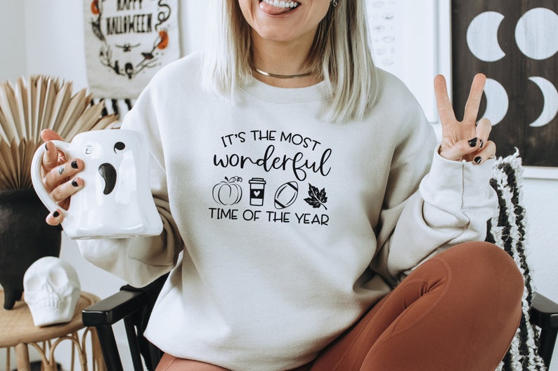 Puede incluir: Sudadera color crema con el texto "It's the most wonderful time of the year" y gr&aacute;ficos tem&aacute;ticos de oto&ntilde;o. El dise&ntilde;o incluye una calabaza, una taza de caf&eacute;, un bal&oacute;n de f&uacute;tbol americano y una hoja. Se sostiene una taza blanca con forma de fantasma.