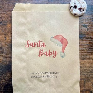 May include: A brown paper bag with the words "Santa Baby" in red script, and a watercolour illustration of a Santa hat. The bag also includes the text "JESSICA'S BABY SHOWER DECEMBER 17TH, 2024". A cookie rests on the bag.