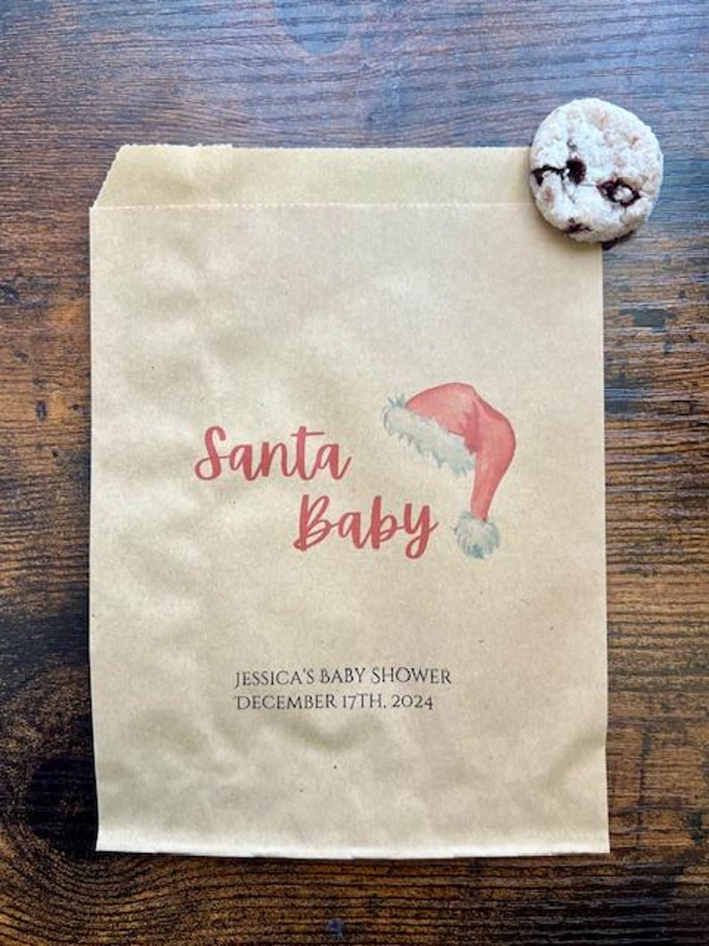 May include: A brown paper bag with the words "Santa Baby" in red script, and a watercolour illustration of a Santa hat. The bag also includes the text "JESSICA'S BABY SHOWER DECEMBER 17TH, 2024". A cookie rests on the bag.