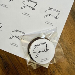 May include: A sheet of circular white adhesive labels featuring the black printed text Midnight Snack and John & Jessica September 5th, 2026. One label is applied to a clear plastic bag containing a dark round cookie with a white cream filling.