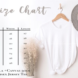 May include: A white Bella + Canvas 3001 Unisex Jersey Tee hanging on a wooden hanger. The image includes a size chart with measurements in inches for XS to 3XL sizes. A gray hat and dried flowers are also visible.