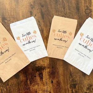 May include: Four paper treat bags with the text "Two little cuties are on the way!" in black and peach-colored script. The bags are in shades of brown and white, perfect for a baby shower.