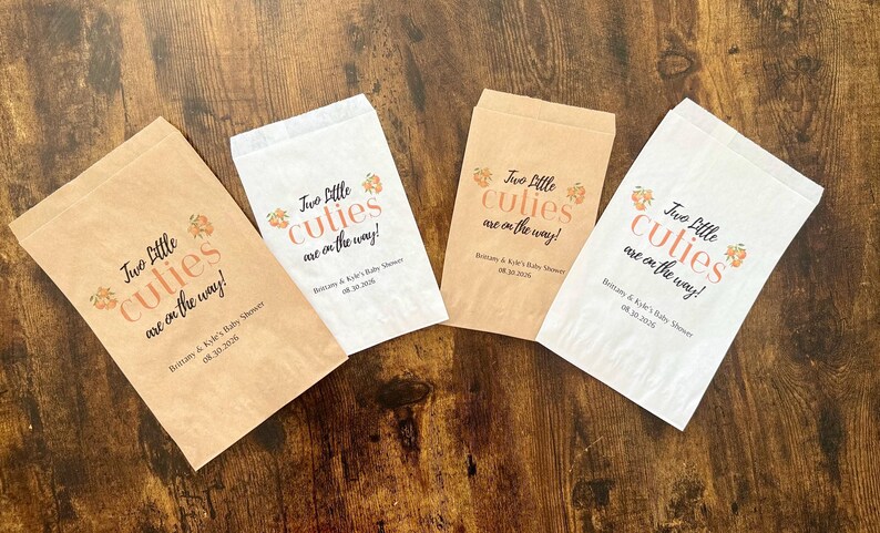 May include: Four paper treat bags with the text "Two little cuties are on the way!" in black and peach-colored script. The bags are in shades of brown and white, perfect for a baby shower.