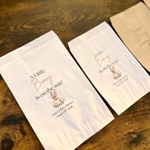 May include: White paper favor bags with the text "A Little Bunny is on the way!" in a script font, along with a watercolor illustration of a bunny. The bags are for Violet's Baby Shower, March 18th, 2026. Some bags are white, others are brown.