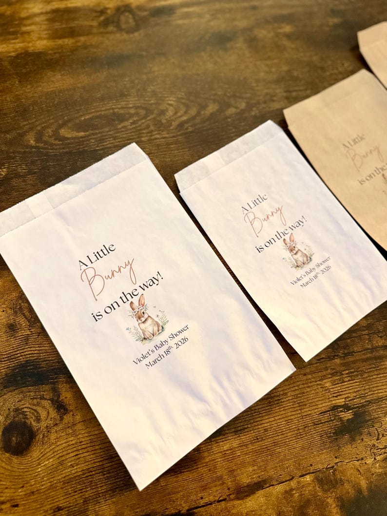 May include: White paper favor bags with the text "A Little Bunny is on the way!" in a script font, along with a watercolor illustration of a bunny. The bags are for Violet's Baby Shower, March 18th, 2026. Some bags are white, others are brown.