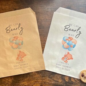 Könnte beinhalten: Zwei Papiertüten, eine beige und eine weiße, mit Babyparty-Motiv. Jede Tüte zeigt einen Teddybären, der Luftballons hält, und den Text "We Can Bearly Wait". Das Datum "April 10, 2024" ist ebenfalls aufgedruckt.