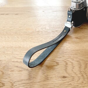 Op de afbeelding: Een zwarte leren camerariem bevestigd aan een zilveren camera. De riem is gelust en heeft een metalen sluiting en klinknagels. De riem ligt op een licht houten oppervlak.