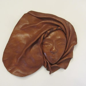 May include: A brown leather wall sculpture featuring a human face partially wrapped in a draped, textured leather fabric. The material has a smooth, polished finish on the facial features and a visible grain pattern throughout the entire piece.