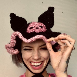 Op de afbeelding: Een zwarte en roze gehaakte muts met varkensoren en een schedelontwerp. De muts heeft een zwart lint dat om de nek is gebonden.