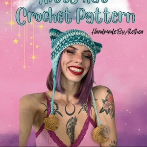 Può includere: Un cappello a forma di gatto in crochet color verde acqua e bianco con orecchie di gatto e un pon pon su ogni orecchio. Il cappello è indossato da una persona con i capelli viola e dei tatuaggi. Lo sfondo è un gradiente rosa e viola con stelle e una luna crescente. Il testo "Night Sky Kitty Hat Crochet Pattern Handmade By Alethea" è visibile.