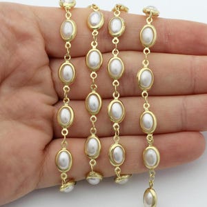 May include: Gold-toned chain featuring oval, faux pearls. Each pearl is set within a gold-colored bezel and linked together with small gold rings. The chain is held against a light background, showcasing the jewelry's design.