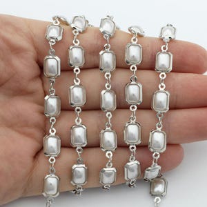 May include: A strand of faux pearl beads with rectangular, silver-toned frames. The pearls are white and the metal has a shiny finish. The beads are connected by small silver links, creating a delicate and elegant design.