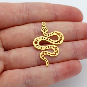 Könnte beinhalten: Ein goldfarbener Schlangenanhänger mit aufwendigen Ausschnitten. Der Anhänger zeigt ein stilisiertes Schlangendesign mit Mond- und Pfeilmuster. Die Schlange ist gewunden und hat oben eine kleine Schlaufe zum Aufhängen. Der Anhänger wird in einer Hand gehalten.