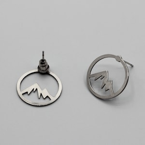 May include: A pair of silver earrings with a mountain range design cut out of the circular shape.