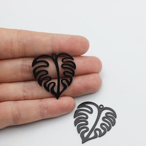 Może przedstawiać: Dwa czarne metalowe charms w kształcie liścia monstera. Charms mają otwór u góry do przymocowania do naszyjnika lub bransoletki.