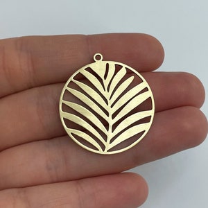 May include: Gold-toned metal pendant with a cutout design of a palm leaf. The pendant is round and has a hole at the top for hanging.