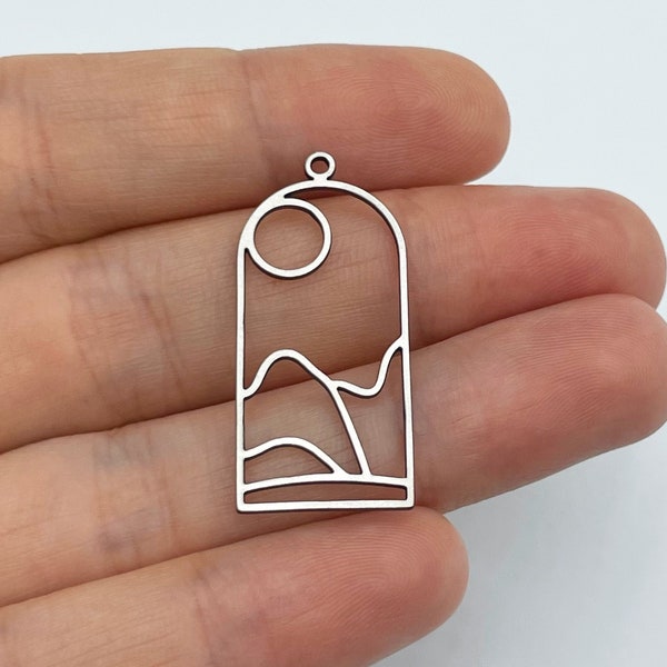 Puede incluir: Pendiente de metal plateado con un diseño geométrico de un sol, montañas y olas. El pendiente tiene un bucle en la parte superior para sujetar una cadena.
