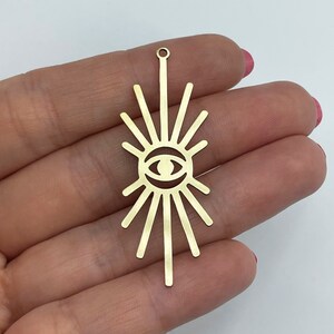Op de afbeelding: Gouden metalen bedel met een zonnestralenontwerp en een oog in het midden. De bedel heeft een gat aan de bovenkant om aan een ketting of armband te bevestigen.