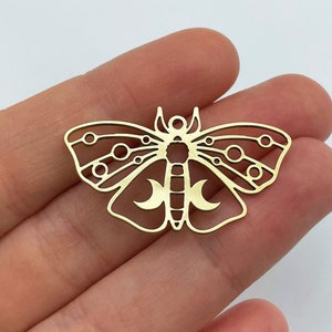 Puede incluir: Un colgante de metal dorado con forma de polilla con intrincados detalles recortados. La polilla presenta formas circulares y de media luna dentro de sus alas y cuerpo. El colgante se sostiene en una mano sobre un fondo blanco.