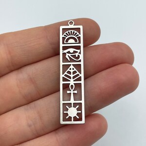 May include: Silver charm with an Egyptian theme. The charm features a sun, an eye of Horus, a tree of life, an ankh, and a sun. The charm is rectangular and has a hole at the top for attaching to a necklace or bracelet.