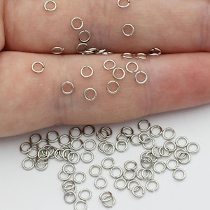 May include: A scattering of small, silver jump rings on a light surface. The round rings are made of metal. Some are held in a hand, while others are loose. These are suitable for jewellery making and crafts. The rings are approximately 0.5 cm in diameter.