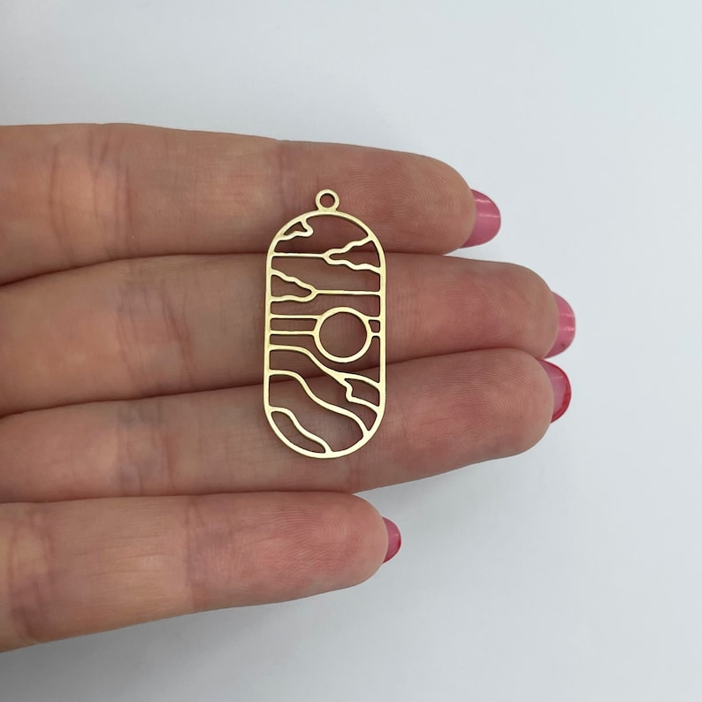 May include: Gold-toned metal pendant with a cutout design of wavy lines and a circle. The pendant is oval-shaped and has a hole at the top for hanging.