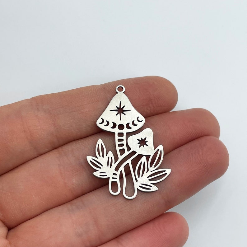 May include: Silver charm featuring a detailed cutout design of two mushrooms with a star and crescent moon details. The charm has a loop at the top for attaching to a necklace or bracelet.