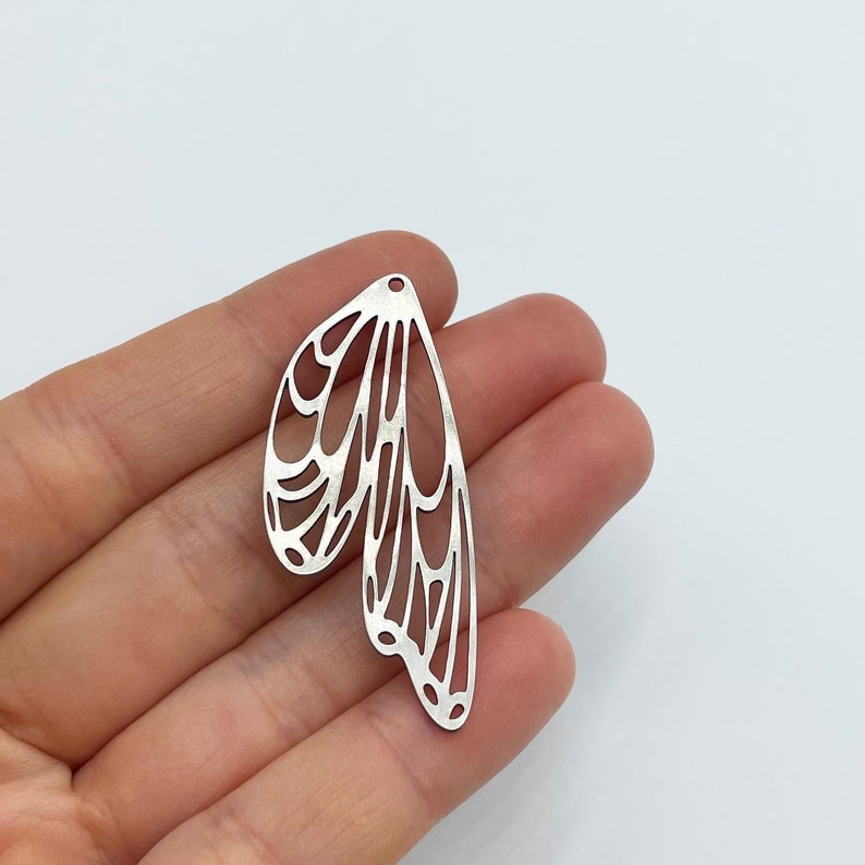 Op de afbeelding: Zilveren metalen bedel in de vorm van een vlinder vleugel met een delicaat, ingewikkeld ontwerp.