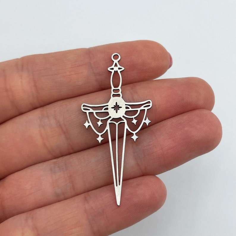 May include: Silver pendant charm in the shape of a dagger with a star in the center and a decorative design around the blade. The pendant is held in a hand.