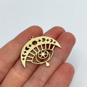 Peut inclure: Breloque en m&eacute;tal dor&eacute; en forme de croissant de lune avec des &eacute;toiles et un &oelig;il stylis&eacute; avec une &eacute;toile &agrave; l'int&eacute;rieur. L'&oelig;il a une forme de larme en bas.
