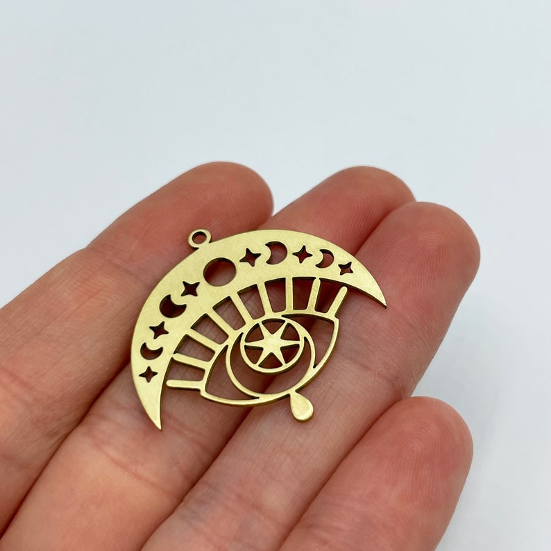 Peut inclure: Breloque en m&eacute;tal dor&eacute; en forme de croissant de lune avec des &eacute;toiles et un &oelig;il stylis&eacute; avec une &eacute;toile &agrave; l'int&eacute;rieur. L'&oelig;il a une forme de larme en bas.