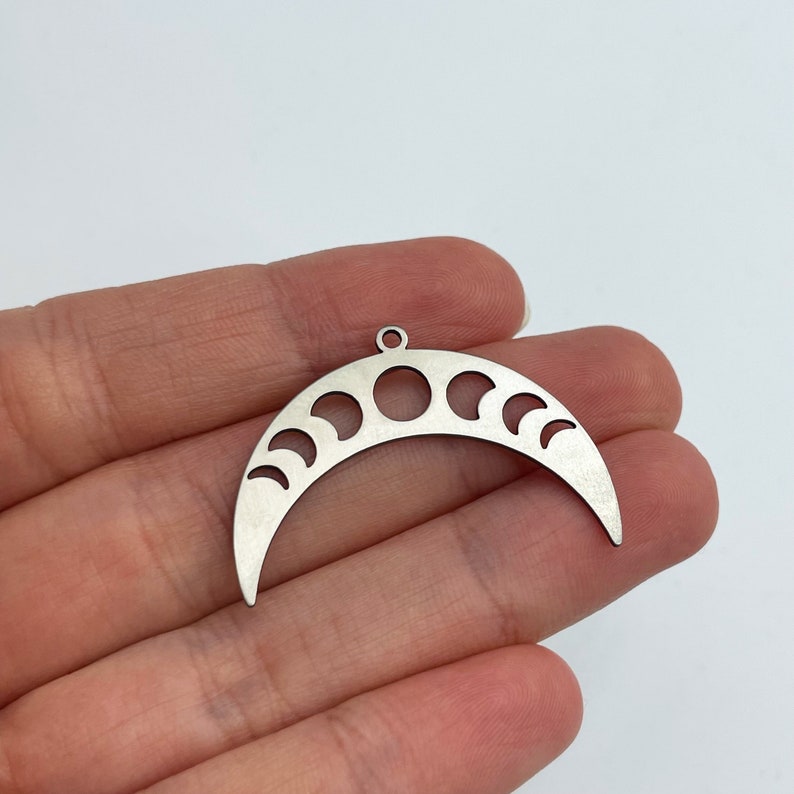 May include: A silver metal crescent moon charm with cut-out phases of the moon. The charm has a loop at the top for attaching to a necklace or bracelet.