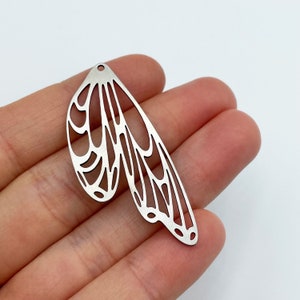 Op de afbeelding: Zilveren metalen bedel in de vorm van een paar vlinder vleugels met een delicaat, ingewikkeld ontwerp.