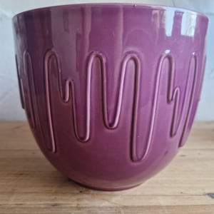 Puede incluir: Una maceta grande y redonda de cerámica morada con un acabado brillante. La maceta presenta una serie de hendiduras verticales y curvas alrededor del exterior. Se encuentra sobre una superficie de madera, adecuada para el hogar o el jardín.