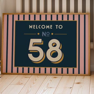 以下が含まれることがあります： レトロなデザインの額入りウォールアート。アートワークは、ヴィンテージフォントで「WELCOME TO NO. 58」というテキストを特徴とし、番号58が大きく表示されています。フレームはピンクとネイビーのストライプのボーダーです。