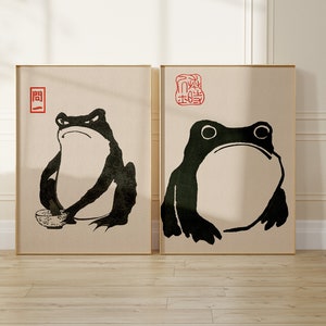 Puede incluir: Dos impresiones enmarcadas de ilustraciones en blanco y negro de ranas. La rana de la izquierda sostiene un cuenco. La rana de la derecha está sentada con la boca abierta. Ambas impresiones tienen caracteres chinos rojos en la esquina superior.