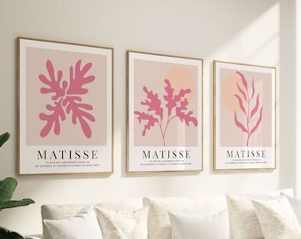 May include: Three framed prints with pink abstract floral designs on a light pink background. Each print has the word "MATISSE" in black text at the bottom. The prints are hanging on a white wall above a white couch with white throw pillows and a white throw blanket.