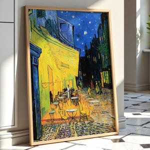 May include: A framed print of Vincent van Gogh's Cafe Terrace at Night, featuring a vibrant yellow cafe exterior illuminated against a deep blue starry sky, with small tables and chairs on a cobblestone street, enclosed in a light wood frame.