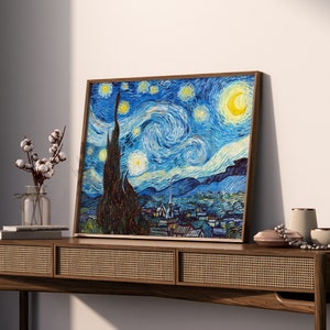May include: A framed painting of a starry night sky with a crescent moon and a swirling sky in shades of blue, yellow, and white. The painting is set against a white wall and a wooden table.