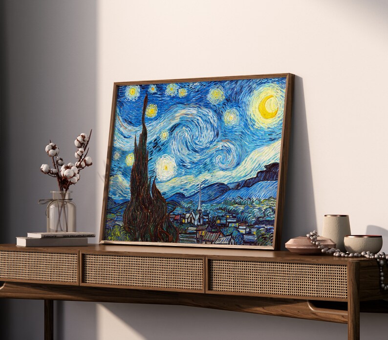May include: A framed painting of a starry night sky with a crescent moon and a swirling sky in shades of blue, yellow, and white. The painting is set against a white wall and a wooden table.