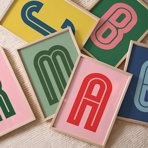 Può includere: Una collezione di stampe artistiche incorniciate con lettere audaci in stile retrò. Ogni stampa presenta una singola lettera in una vivace tavolozza di colori, tra cui rosa, verde, giallo e blu. Le lettere hanno un design geometrico arrotondato, su sfondi a tinta unita.