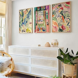 May include: Three framed abstract paintings in a modern living room. The paintings are in a variety of colours, including pink, blue, green, and yellow. The paintings are hung on a white wall above a white dresser with wicker drawers. There are plants in pots on the floor in front of the dresser.