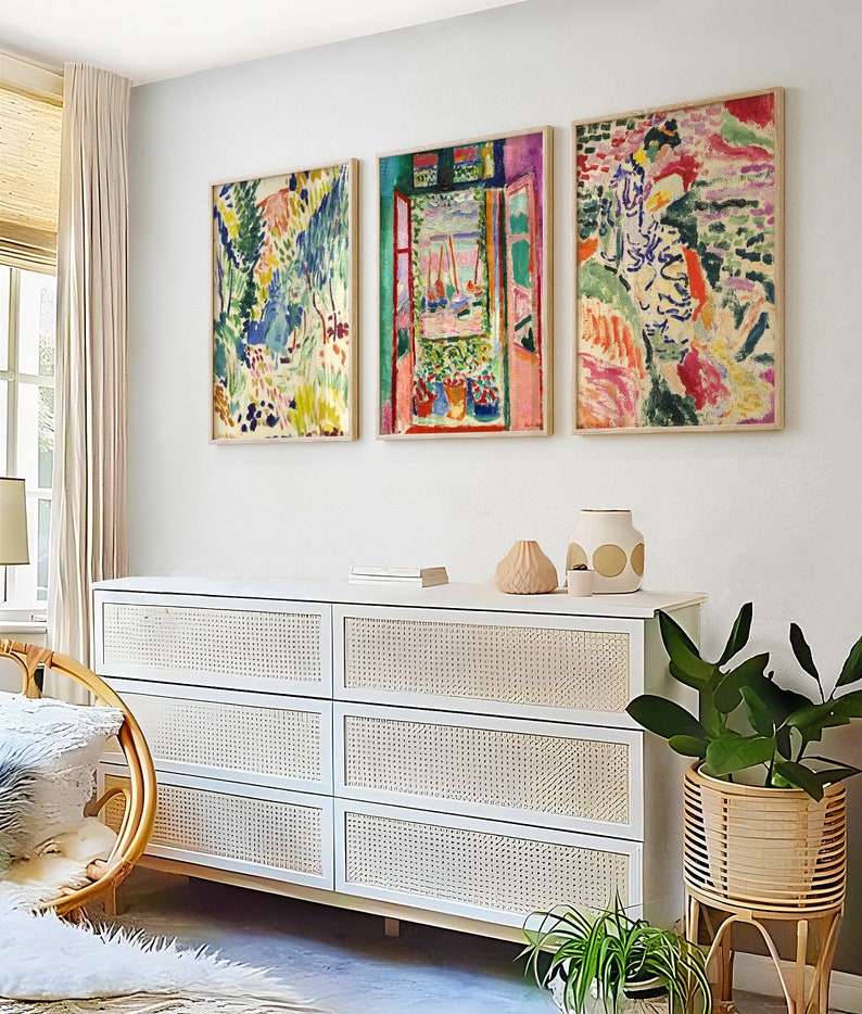 May include: Three framed abstract paintings in a modern living room. The paintings are in a variety of colours, including pink, blue, green, and yellow. The paintings are hung on a white wall above a white dresser with wicker drawers. There are plants in pots on the floor in front of the dresser.