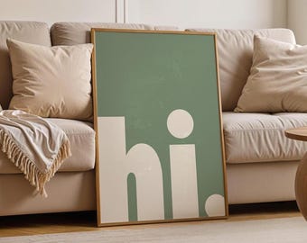 May include: A framed art print featuring the word "hi." in white against a muted green background. The artwork has a minimalist design with a wooden frame, suitable for home decor. The print is leaning against a beige sofa.