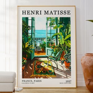Puede incluir: Una impresión artística enmarcada que presenta una escena interior vibrante con una puerta abierta que da al mar. La impresión, etiquetada como "HENRI MATISSE", incluye el texto "FRANCE, PARIS 1937". La obra de arte muestra exuberante vegetación, una alfombra estampada y una silla.