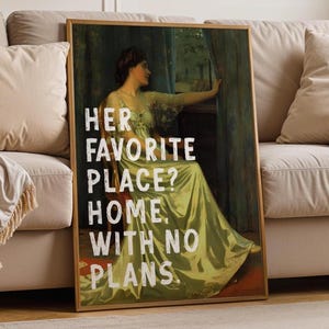 Op de afbeelding: Een ingelijste kunstdruk met een vintage schilderij van een vrouw in een groene jurk. De tekst "HER FAVORITE PLACE? HOME, WITH NO PLANS" is in het wit overgelegd. Het kunstwerk wordt op een beige bank weergegeven.
