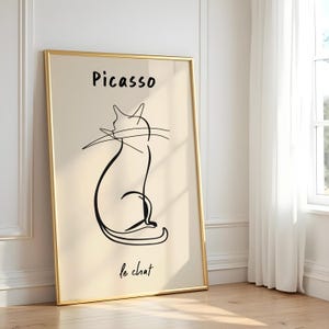 Könnte beinhalten: Ein gerahmter Kunstdruck mit einer minimalistischen schwarzen Strichzeichnung einer Katze auf beigem Hintergrund. Der Druck trägt die Aufschrift "Picasso" über der Katze und "le chat" darunter. Der Rahmen ist goldfarben.