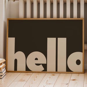 Puede incluir: Un cuadro enmarcado con la palabra "hello" en letras grandes y blancas sobre un fondo marrón oscuro. El marco es de madera clara y la obra de arte se exhibe sobre un suelo de madera.