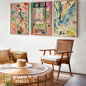 May include: Three framed abstract paintings with colourful brushstrokes depicting a woman, a window with a view of a harbour, and a landscape with trees. The paintings are hanging on a white wall above a wicker coffee table and a wooden chair.